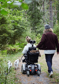 Två personer på hundpromenad i skogen. En av personerna använder permobil.
