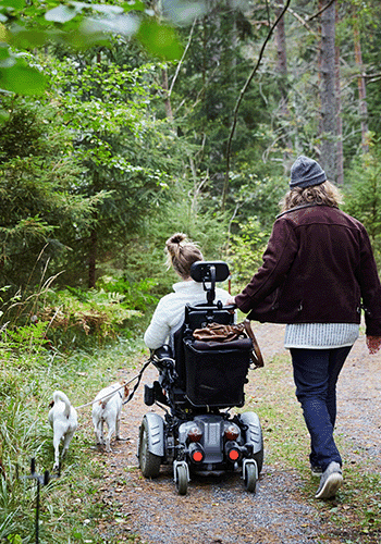 Två personer på hundpromenad i skogen. En av personerna använder permobil.