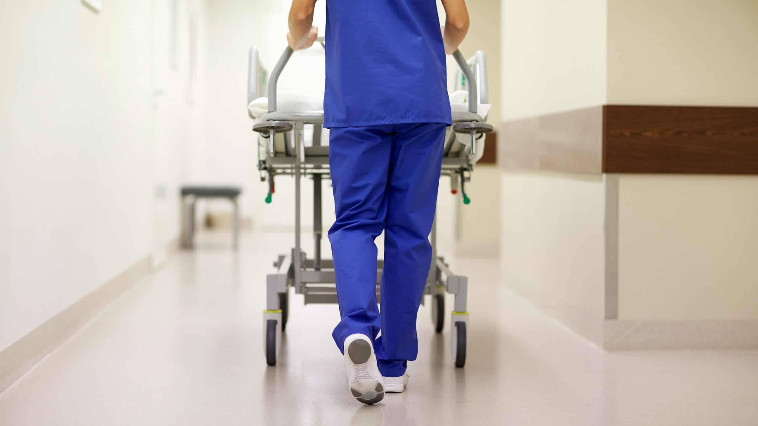 En vårdpersonal i blå kläder skjuter en sjukhussäng framför sig.